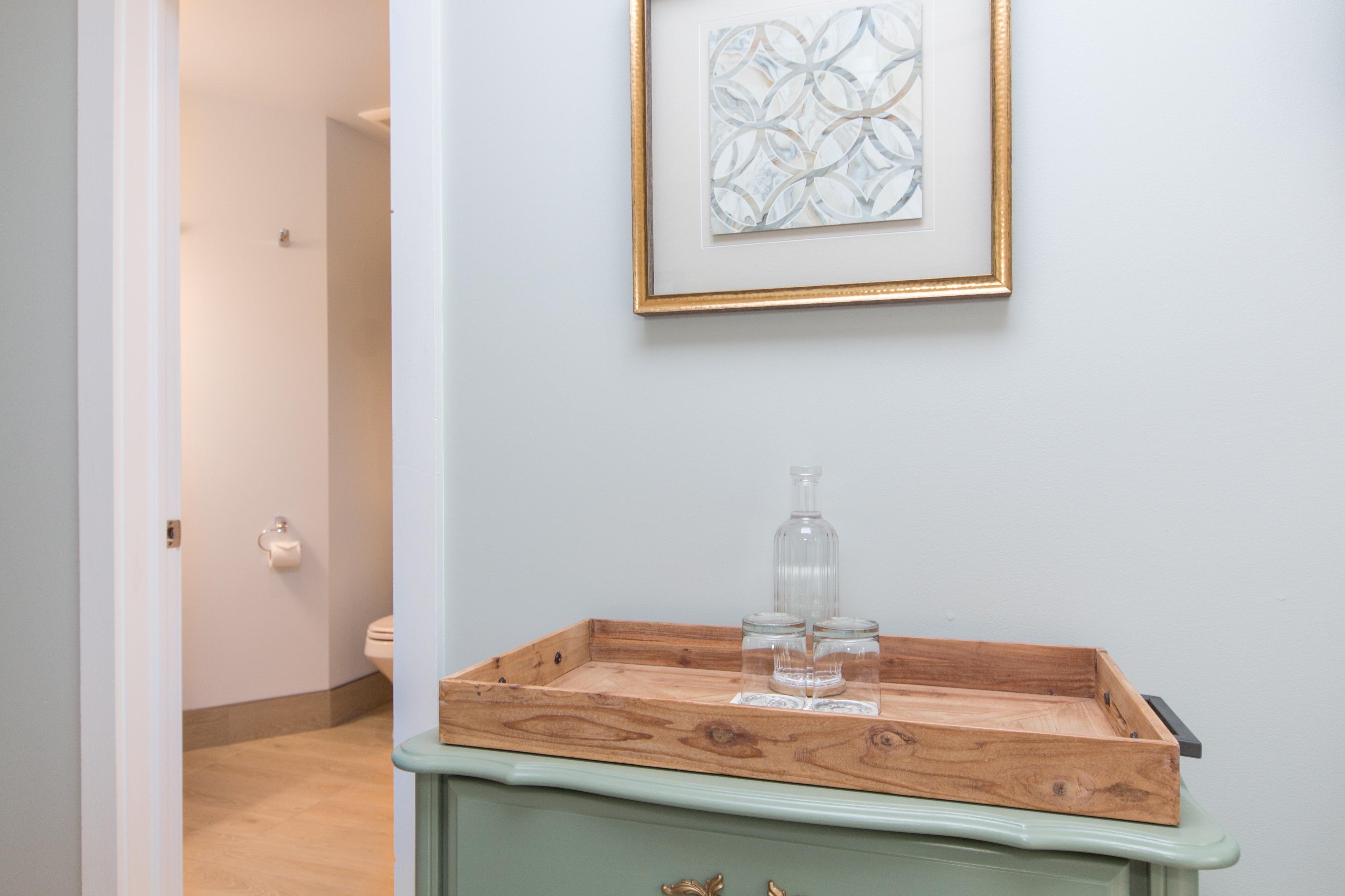 A wooden tray with a glass water bottle sits on a wooden serving tray on top of a green dresser next to an open door leading to an ensuite bath.