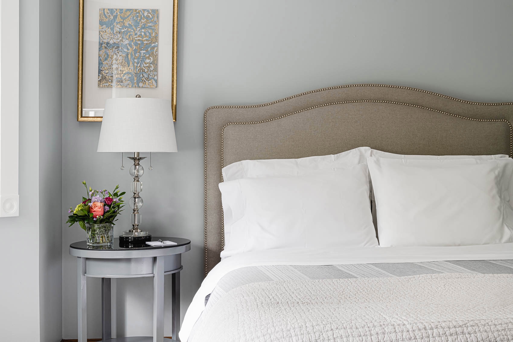 A neatly made bed with white sheets and pillows, a bedside table with a lamp and flowers, and a framed picture on the wall.