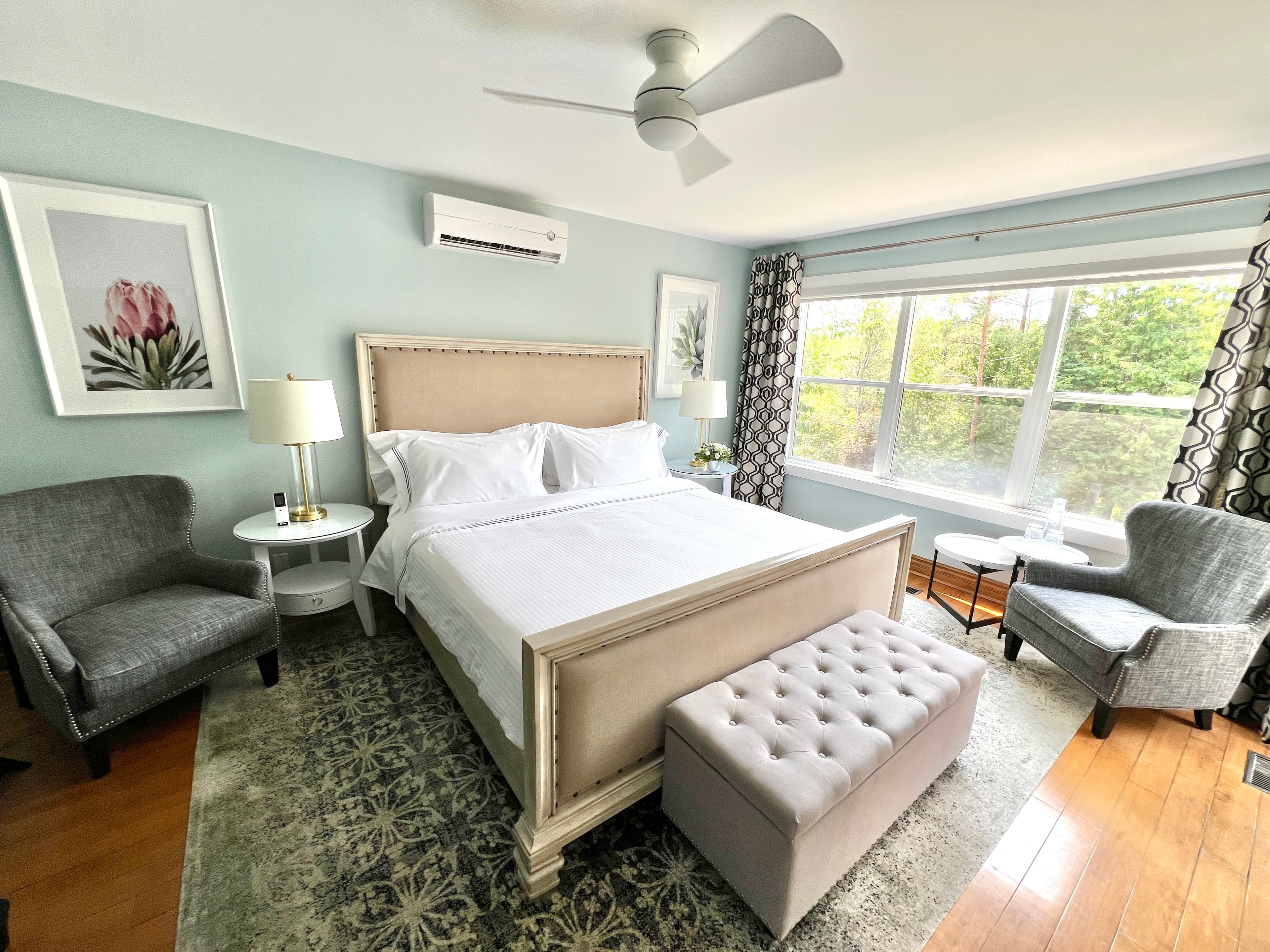 A well-lit bedroom with a king-size bed, two armchairs, a large window with curtains, and a ceiling fan. The walls are painted a light blue color.