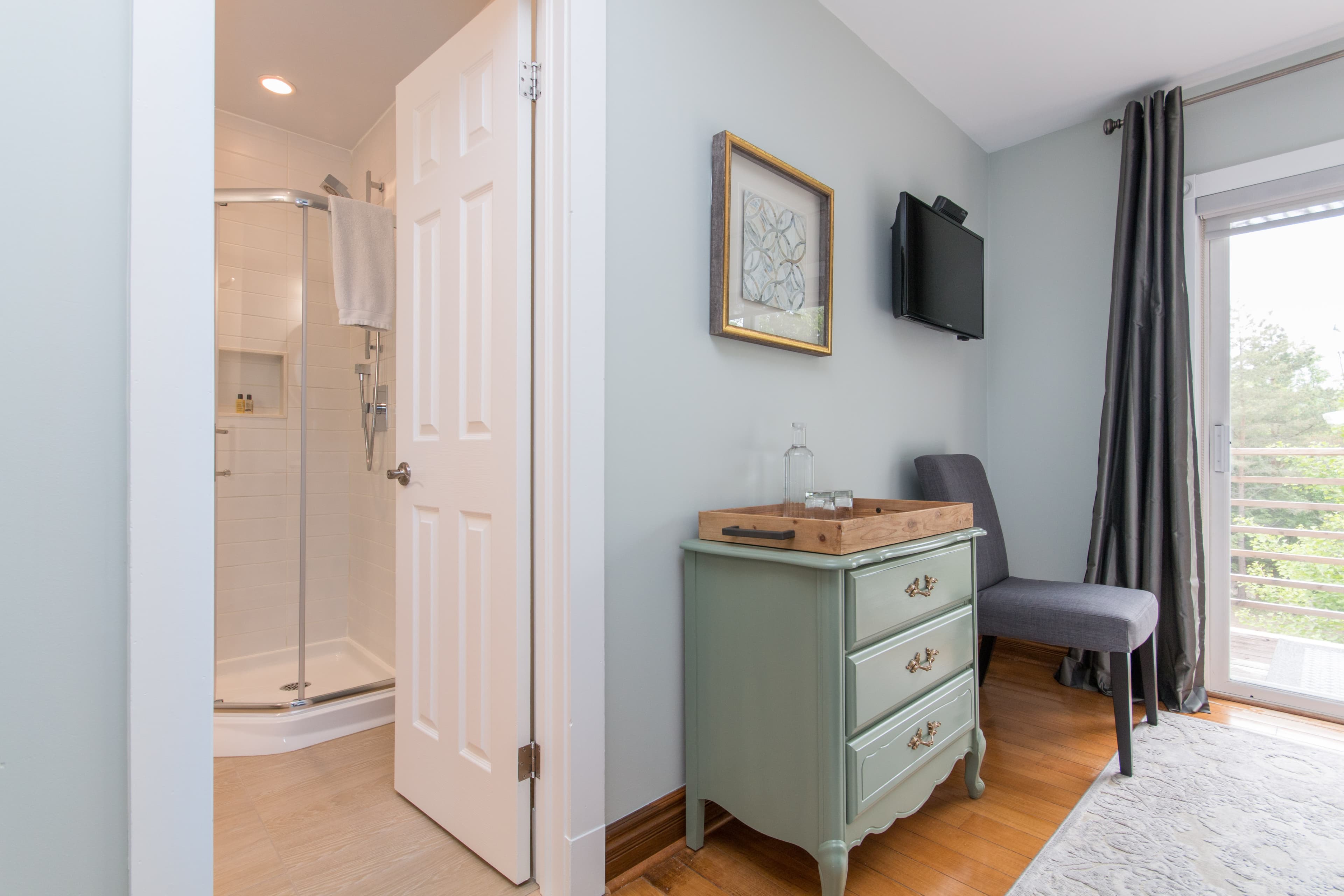 An open door from the bedroom reveals a bathroom with a shower; next to the door in the bedroom is a chair, a dresser, and a television.