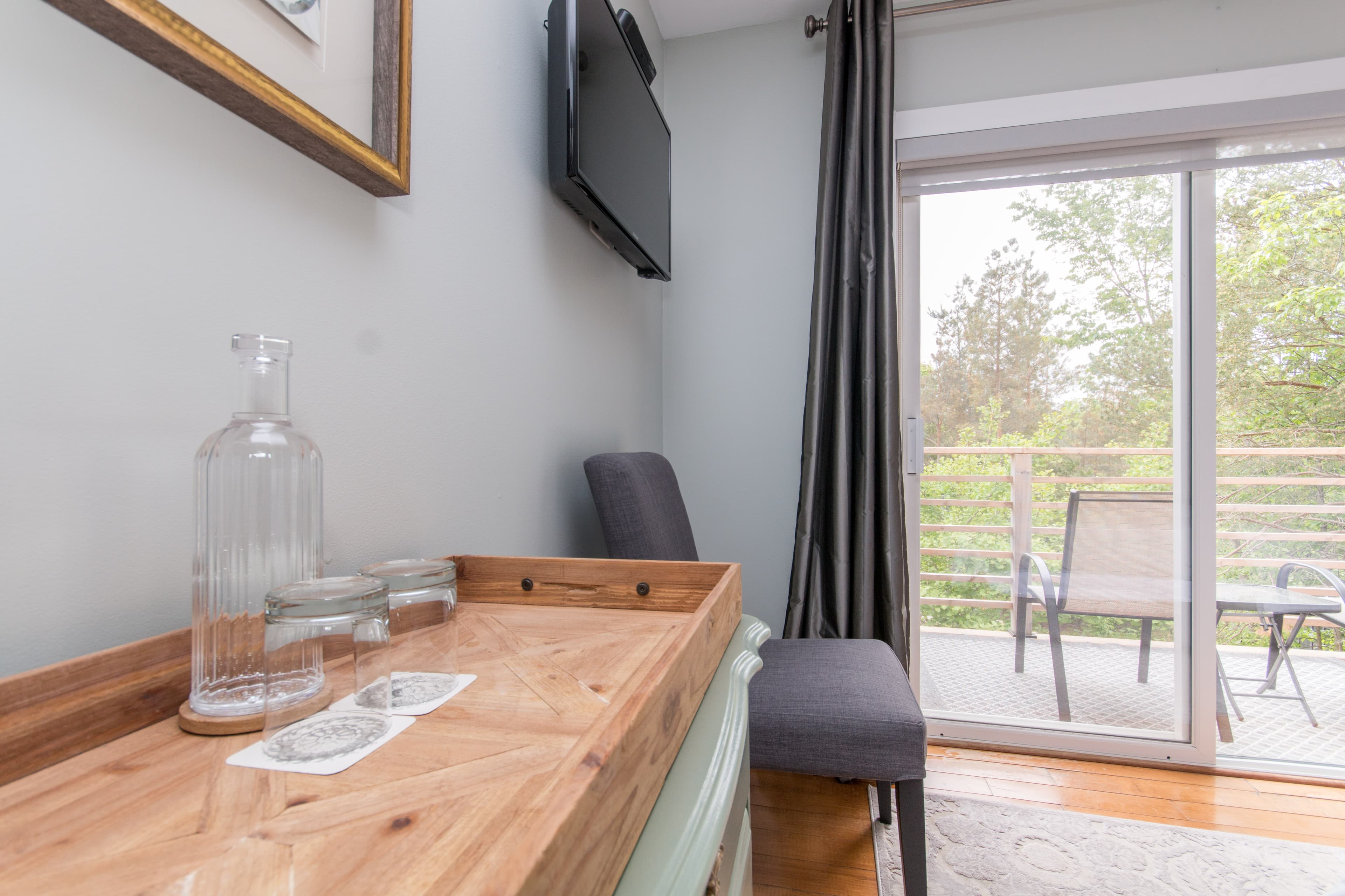 A wooden tray with a glass water bottle and glasses on a dresser next to a chair. A television is mounted on the wall, and a sliding glass door leads to a private balcony with a view of trees.