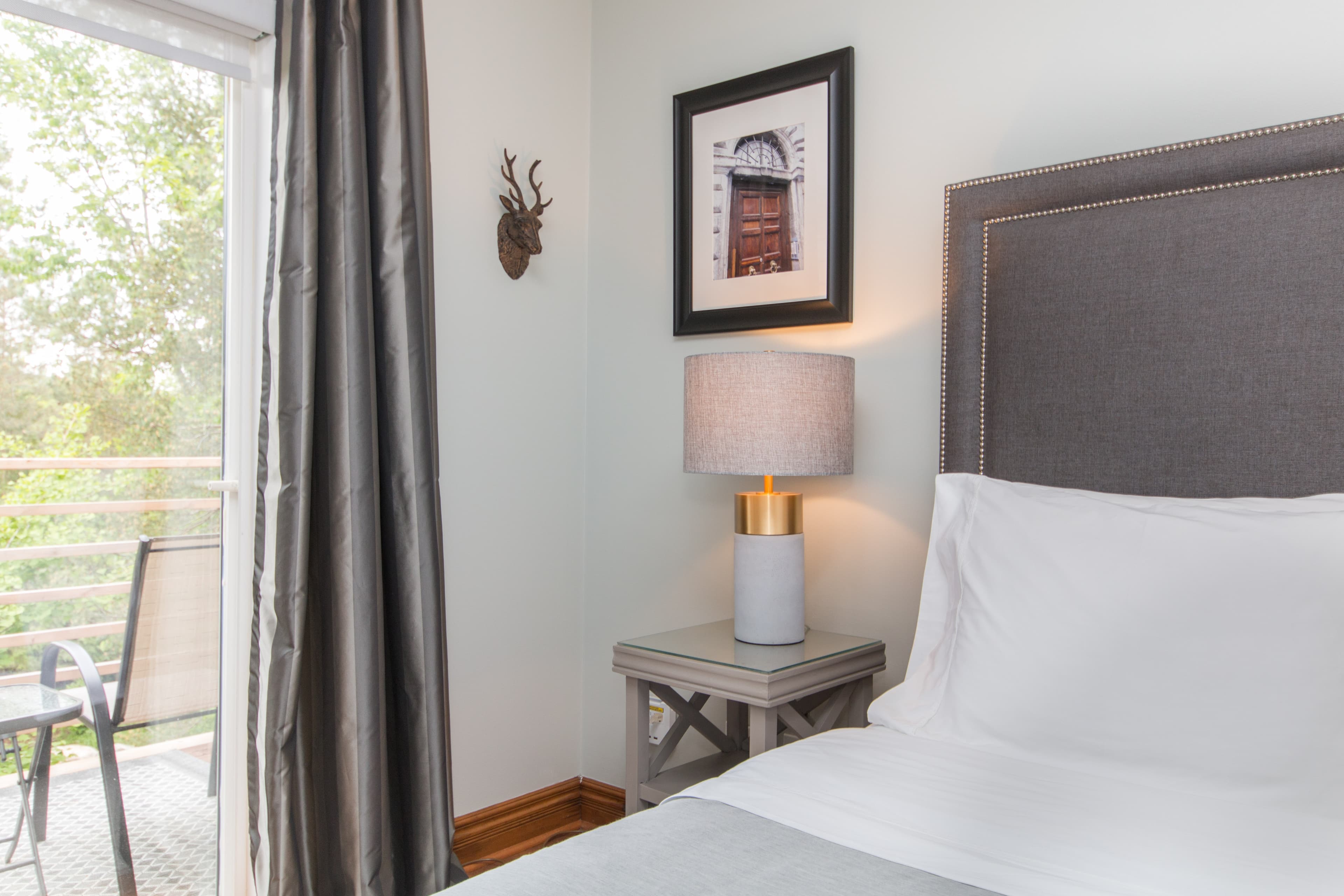 A bedroom with a bed, a nightstand with a lamp, a framed picture, a deer sculpture, and a sliding glass door leading to a balcony with a view of trees.