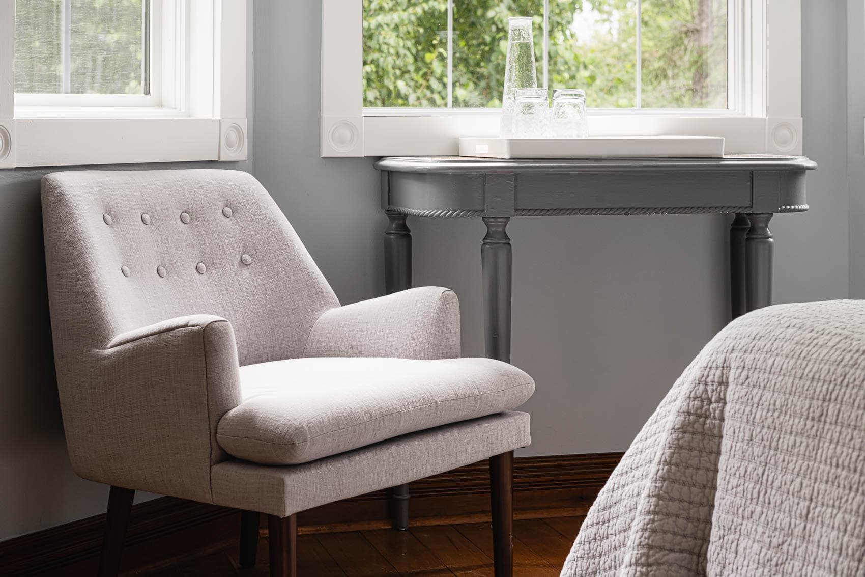 A cozy corner of a bedroom with a white armchair, a small table, and a bed. The window has curtains and a view of trees.