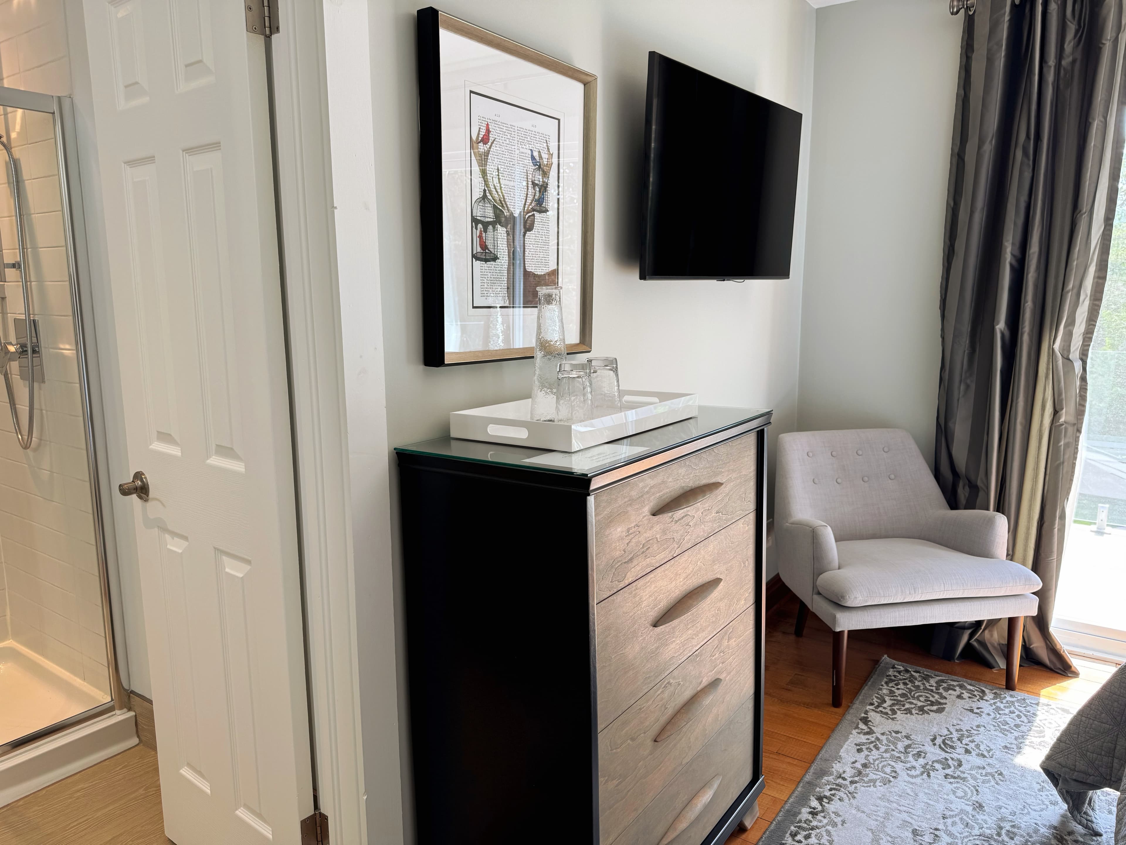 An open door from the bedroom reveals a bathroom with a shower; next to the door in the bedroom is a chair, a dresser, and a television.