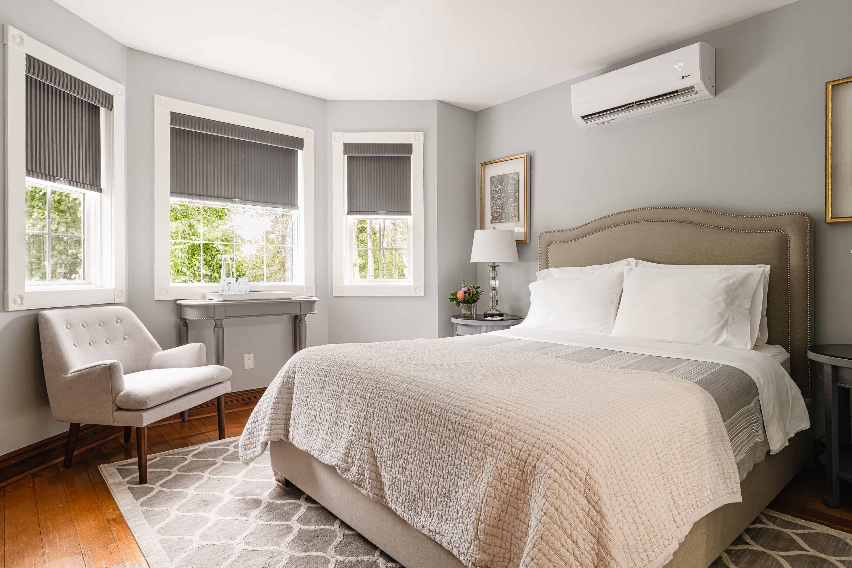A spacious bedroom decorated in shades of taupe, grey, and white with a queen-sized bed, a large window with curtains, a chair, a side table, and a ceiling fan.