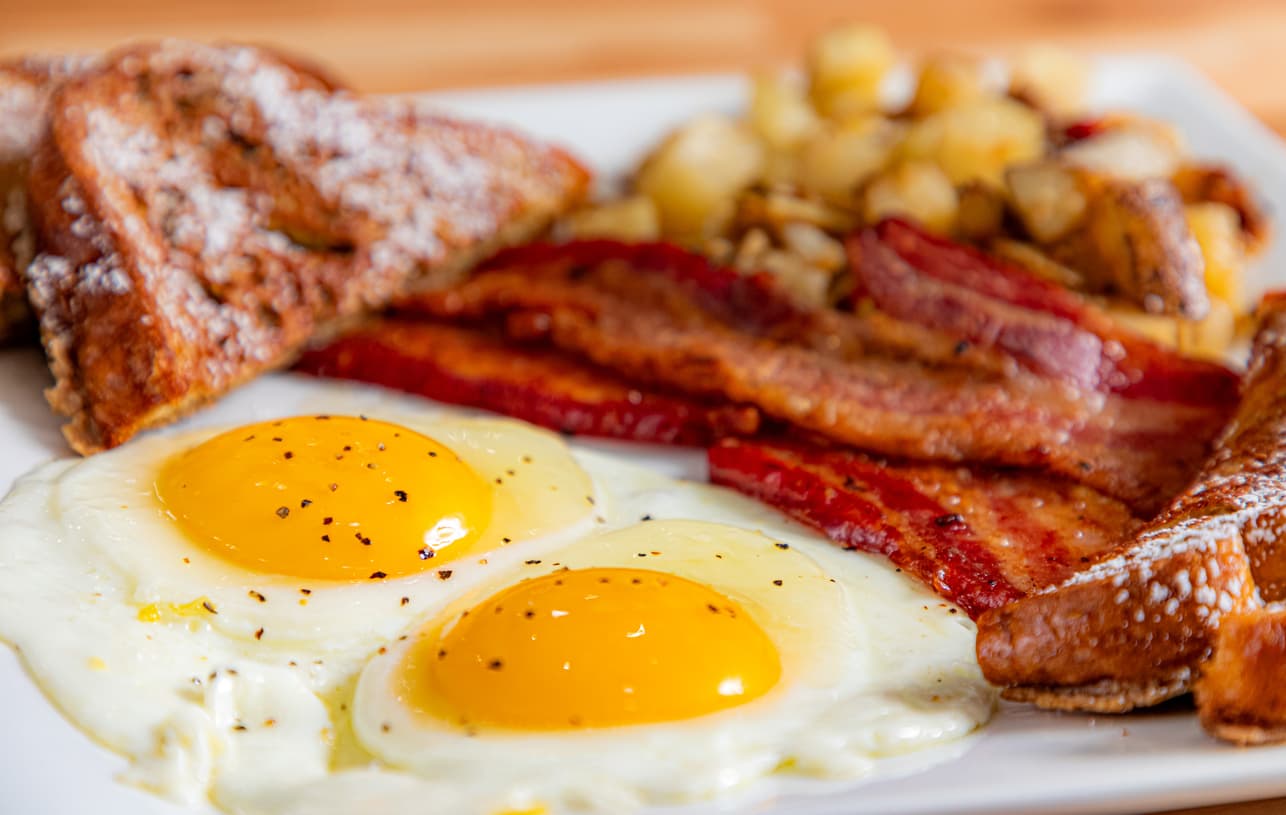 A plate of breakfast featuring two sunny-side-up eggs, crispy bacon, French toast, and seasoned potatoes.