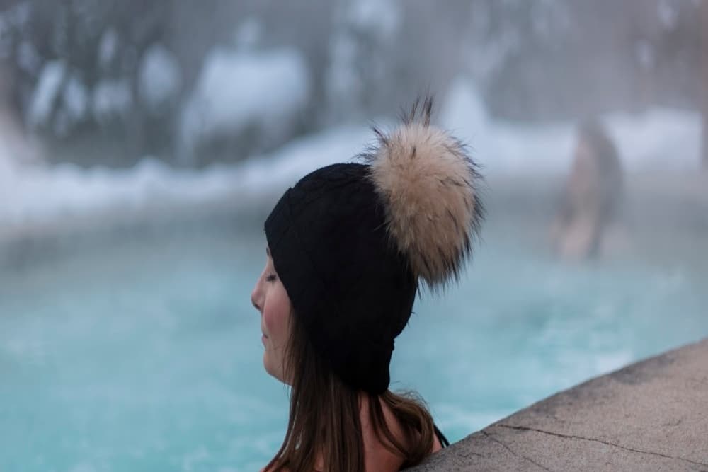 A woman in a black beanie with a fur pom-pom relaxes in a steaming hot spring.
