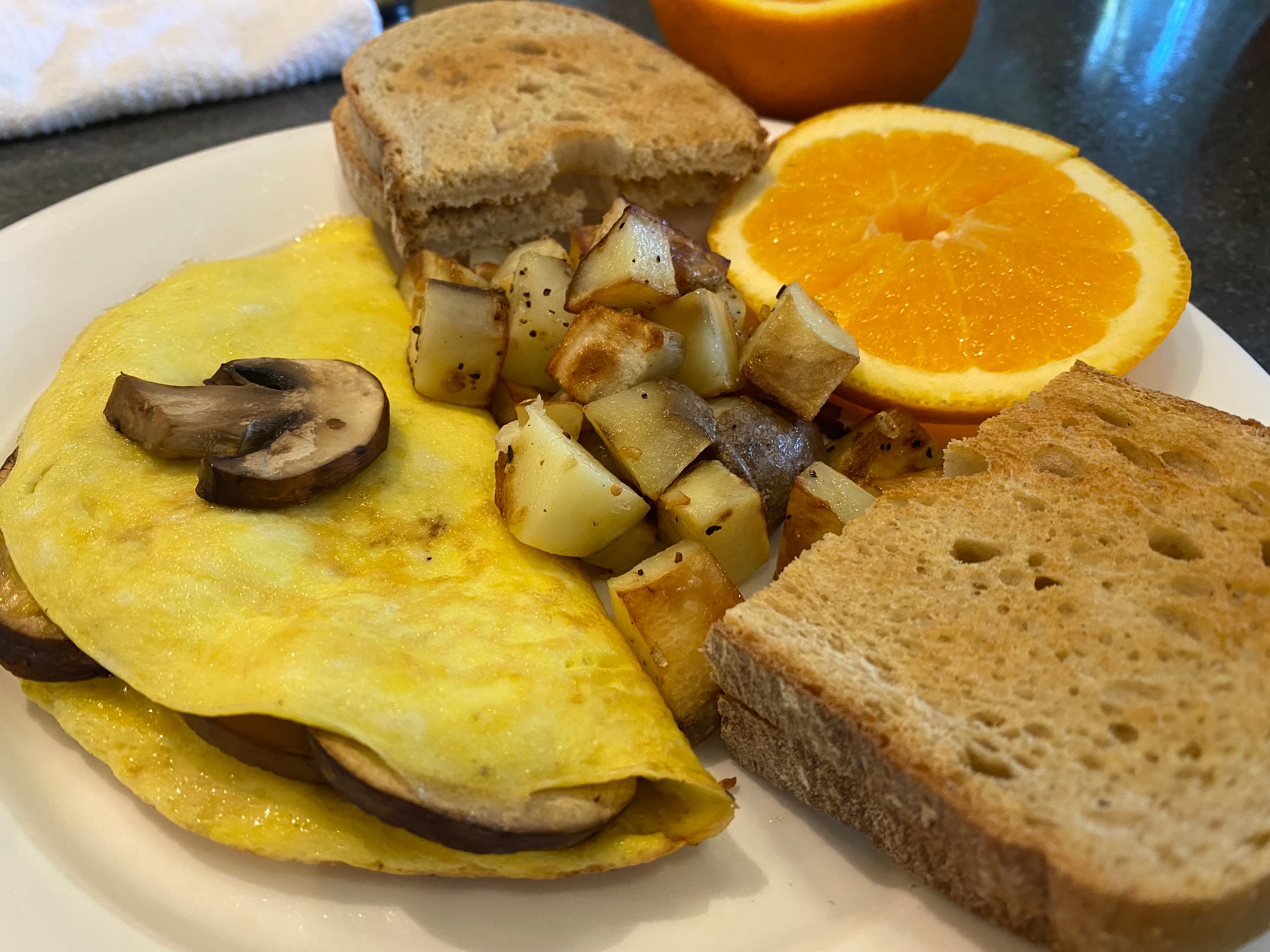 A plate of breakfast featuring a mushroom omelet, roasted potatoes, whole grain toast, and a halved orange.