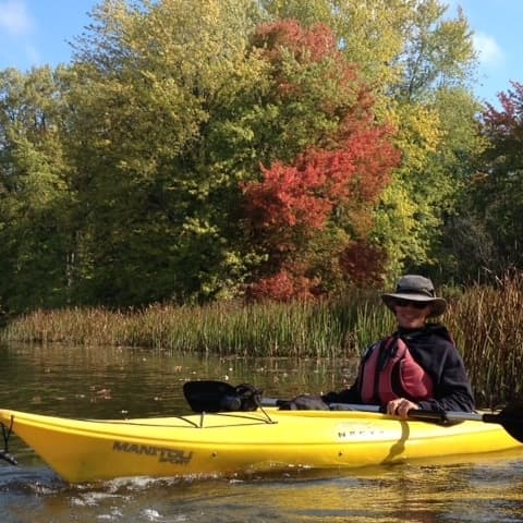 A person in a yellow kayak paddles on a calm body of water surrounded by vibrant autumn foliage.