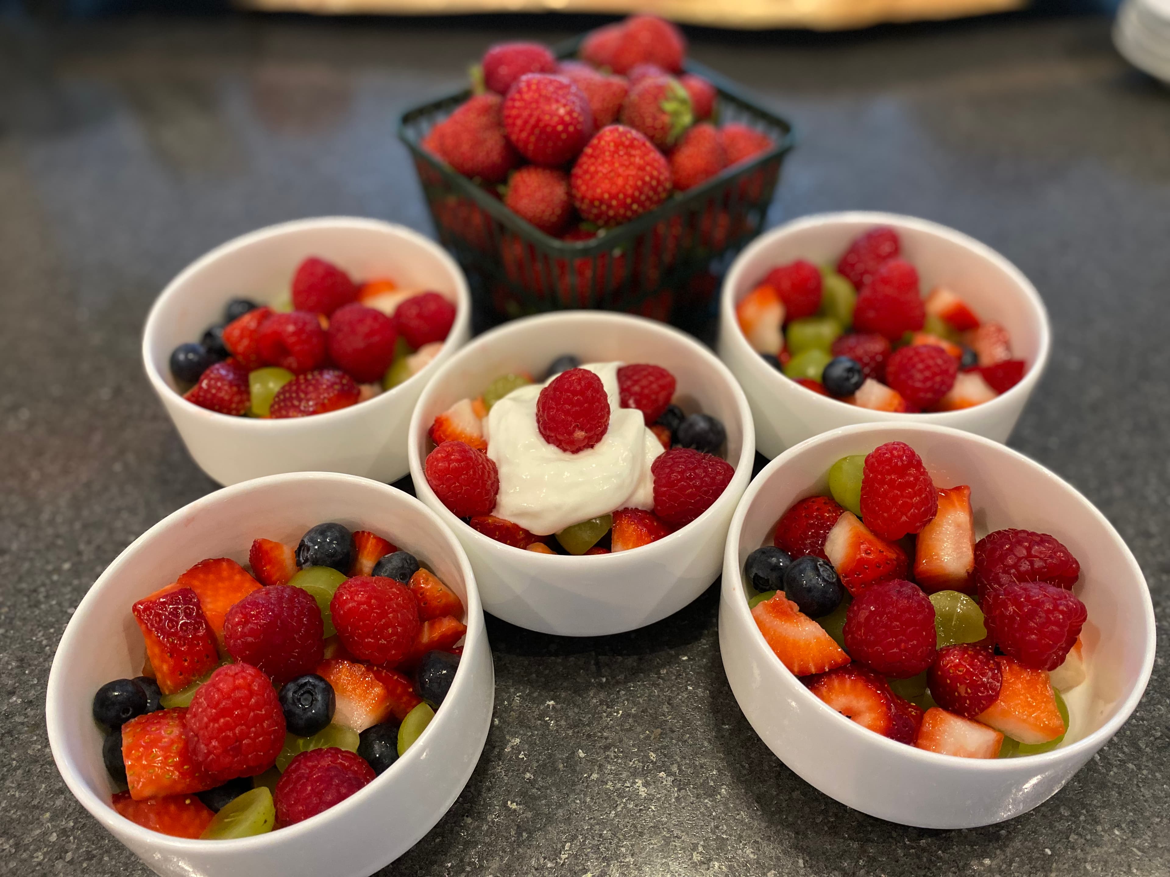Five bowls of mixed fruits topped with raspberries, blueberries, and grapes, with a basket of strawberries in the background.