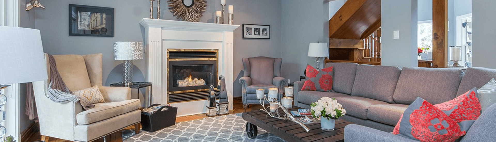 Grey couch red pillows, coffee table, tan chairs in front of fireplace with lamps and free standing lighting nearby