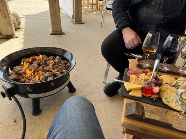 A fire pit with sizzling meat sits next to a wooden platter of cheese and charcuterie, accompanied by drinks.