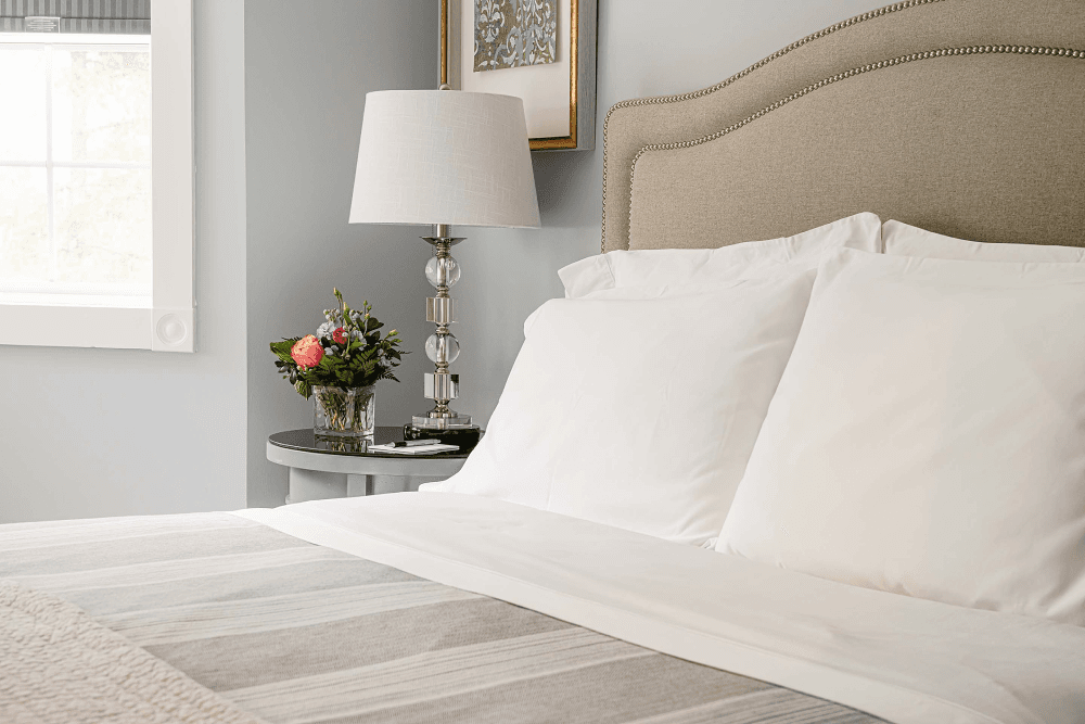 A cozy bedroom featuring a neatly made bed with white pillows, a bedside table with a lamp, and a floral arrangement.