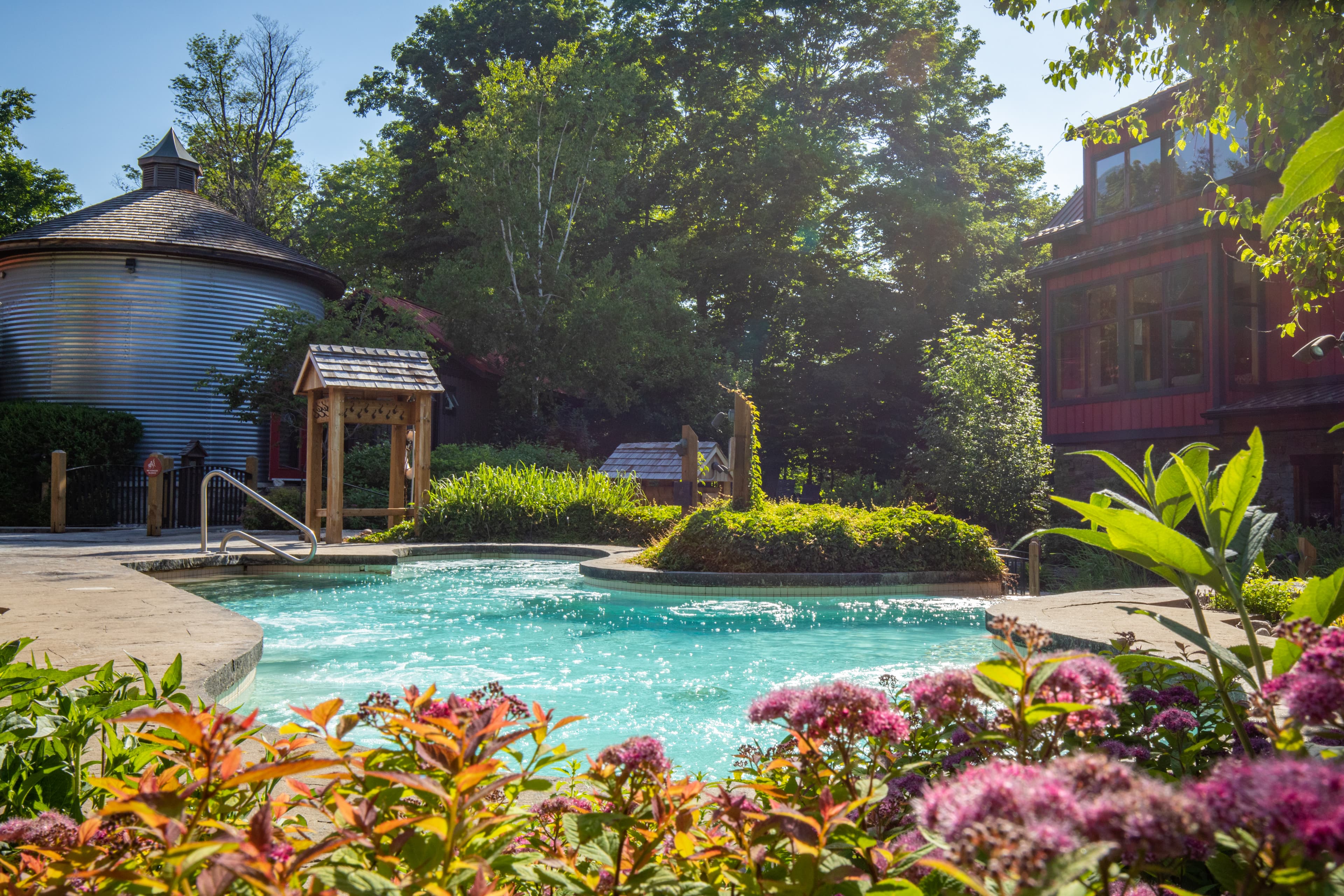 A tranquil pool surrounded by greenery and colorful flowers, with rustic buildings in the background.