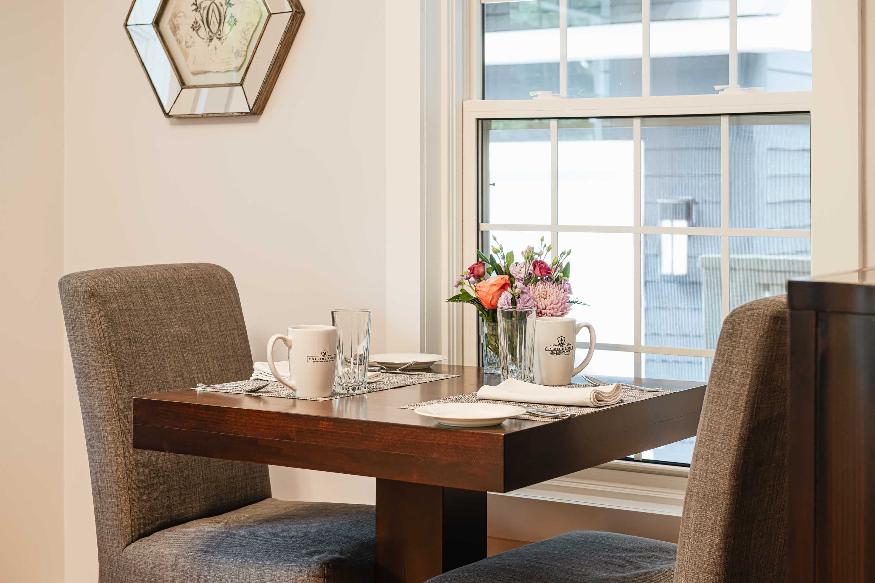 A cozy dining table set for two, featuring mugs, a glass, and a vase of flowers by a window.
