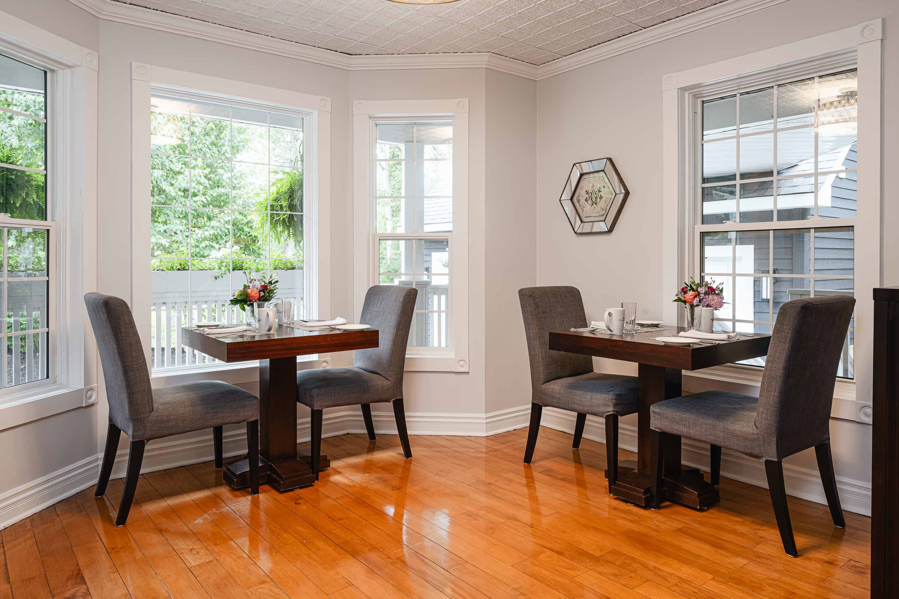 A cozy dining area with two tables, gray chairs, and sunlight streaming in through large windows.