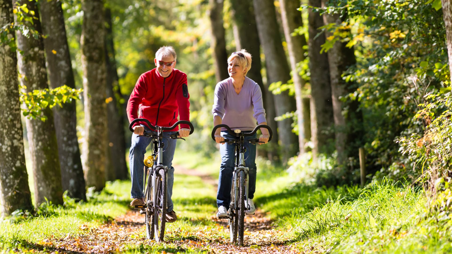 Two elderly individuals enjoy cycling together on a lush, tree-lined path.
