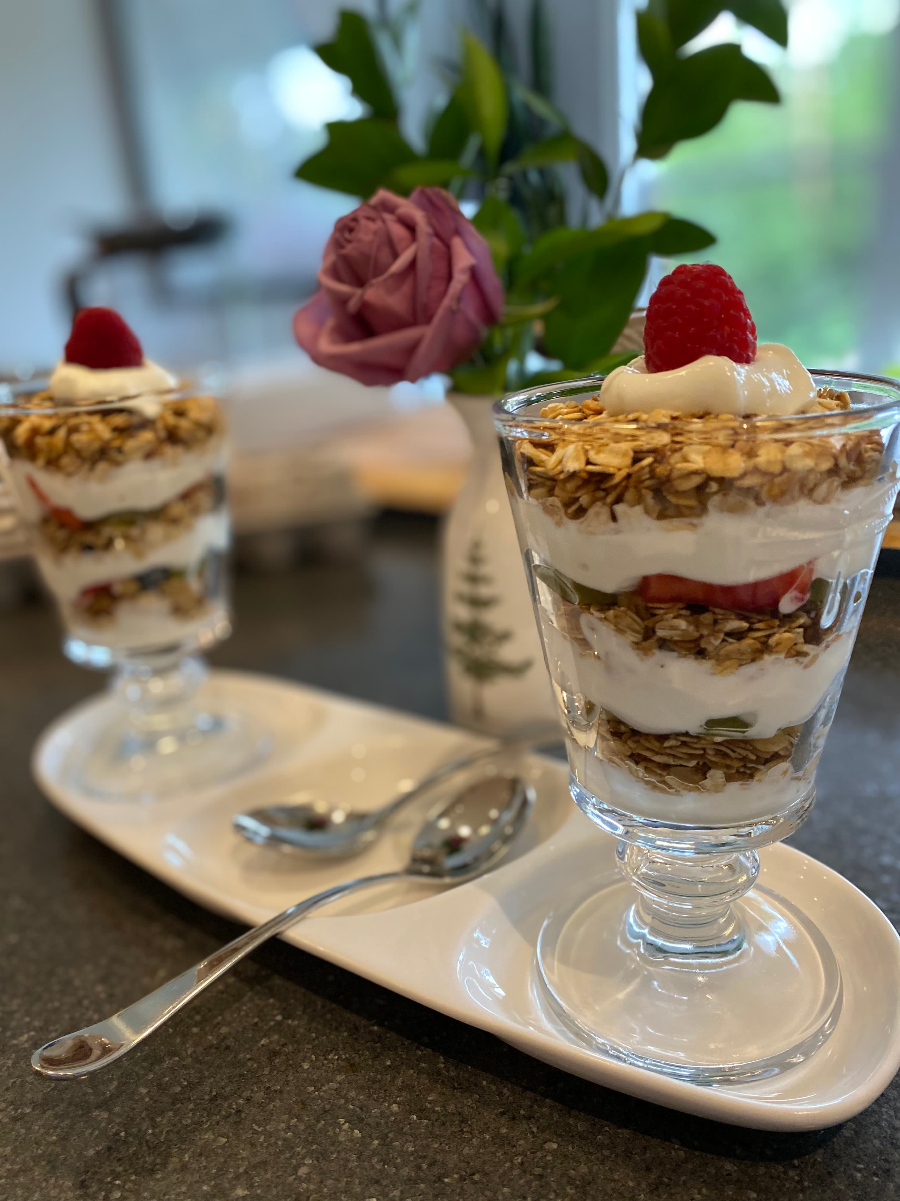 Two layered parfaits with granola, yogurt, and raspberries sit on a white platter next to a rose in a vase.