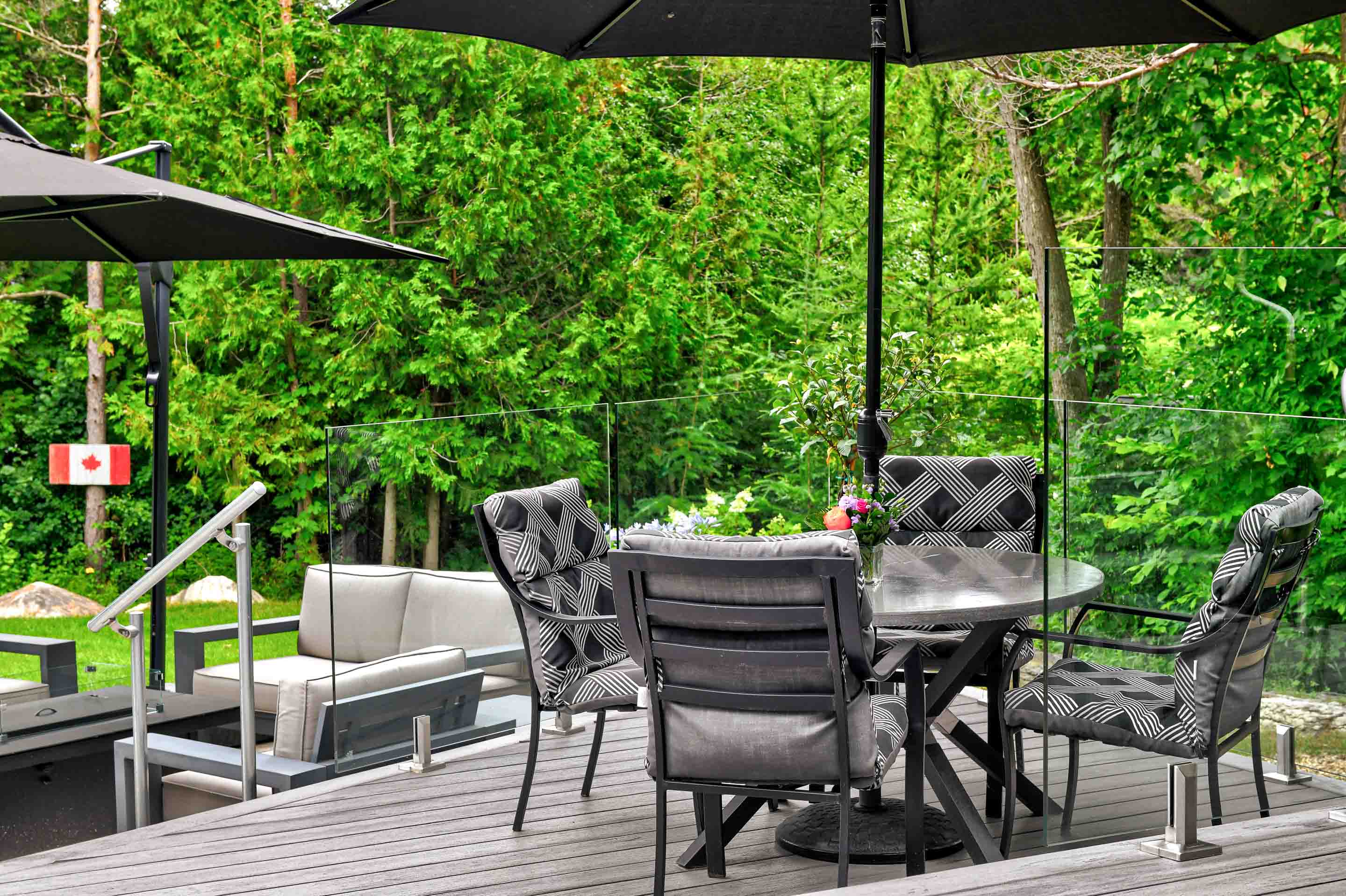A outdoor dining area with black chairs and a round table, surrounded by greenery and a Canadian flag in the background.