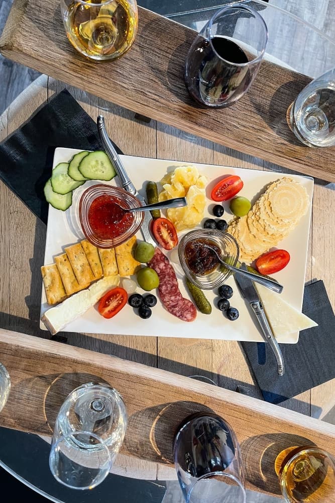 A cheese and charcuterie board featuring assorted crackers, fruits, and condiments, surrounded by glasses of different drinks.