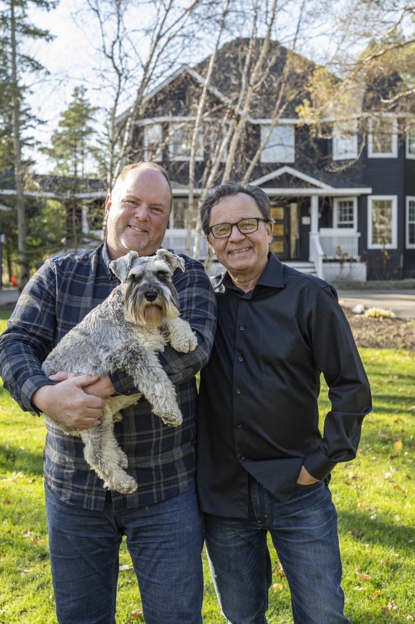 Two men smile together in a grassy area, one holding a dog, with a scenic house in the background.