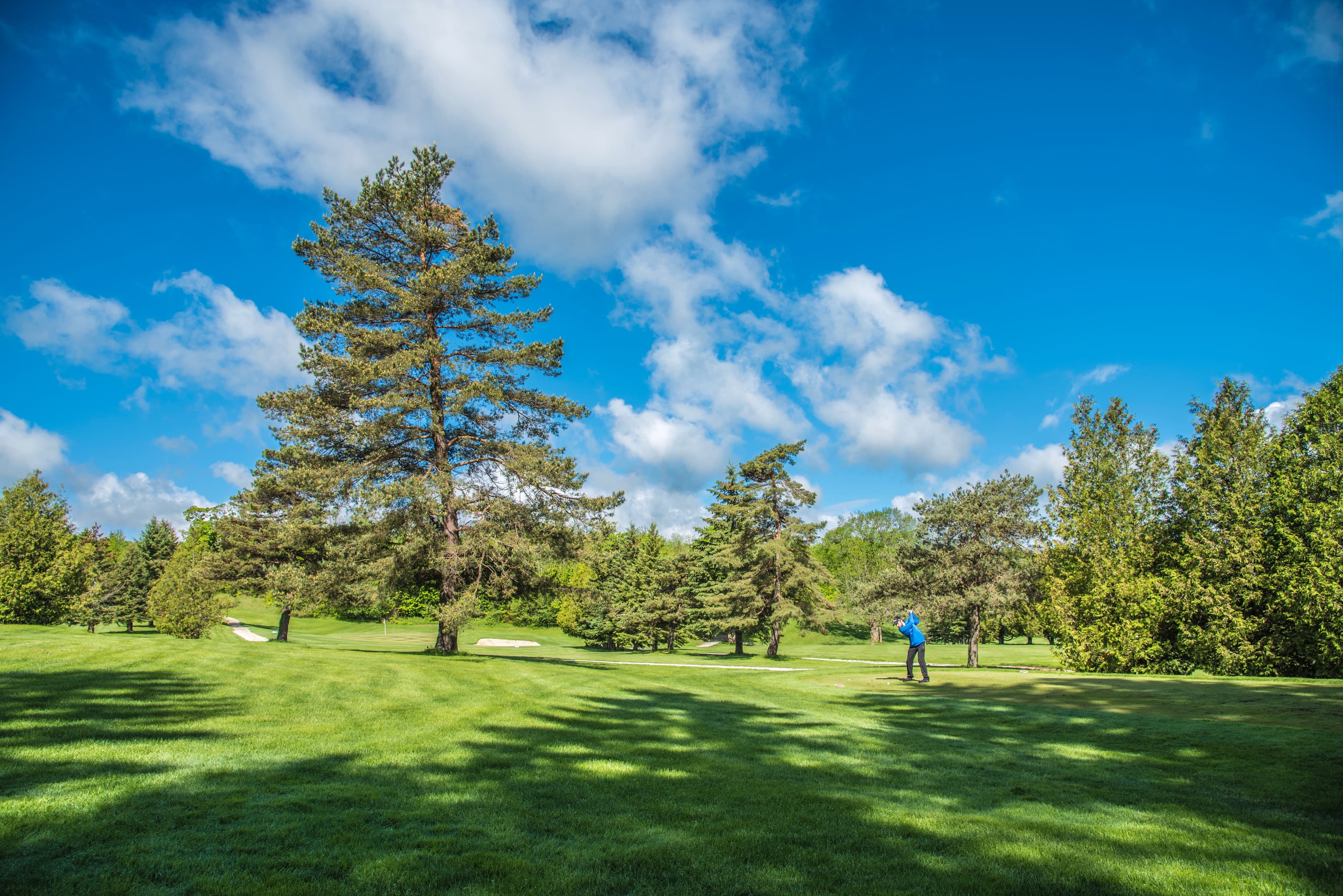A golfer swings on a lush green course under a blue sky with scattered clouds.
