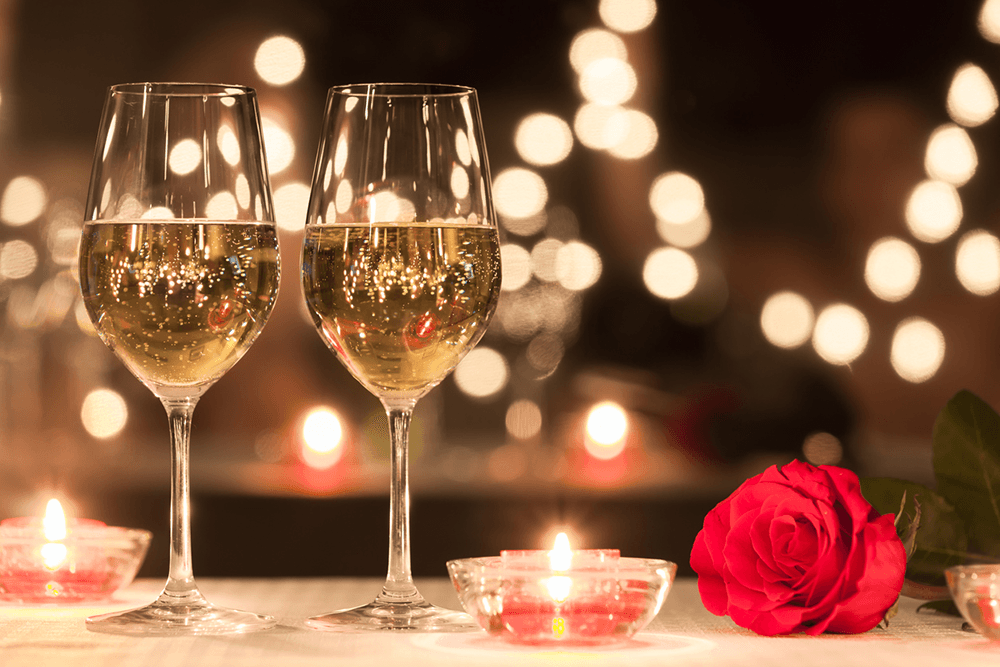 Two glasses of white wine beside candles and a red rose, against a softly lit background.