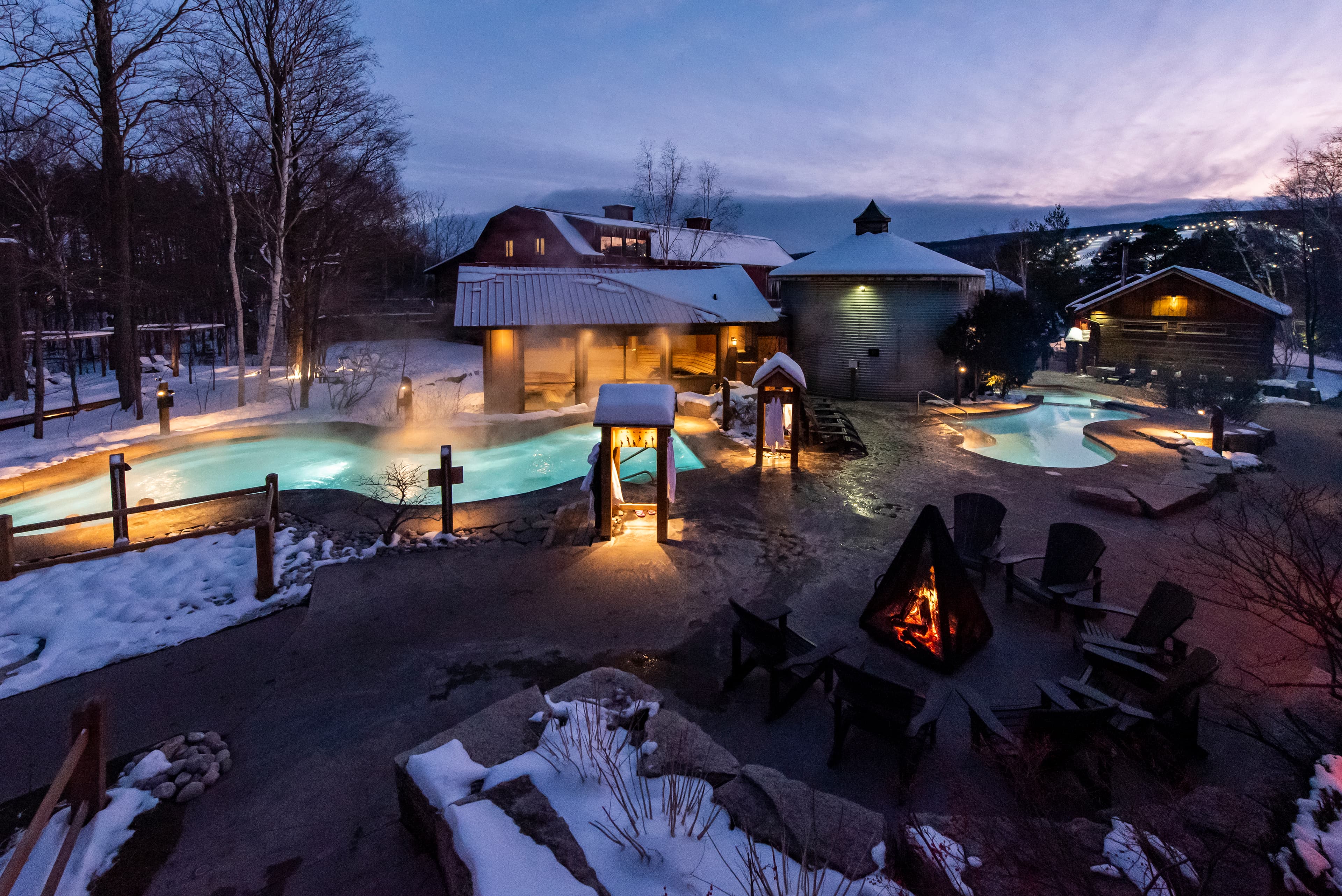 A dusk view of an outdoor hot spring area surrounded by snow, featuring glowing pools and a campfire.