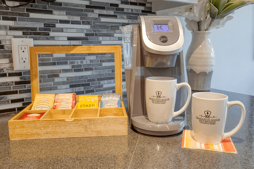 A coffee station featuring a Keurig machine, a bamboo tea box filled with assorted tea bags, and two branded mugs on a countertop.