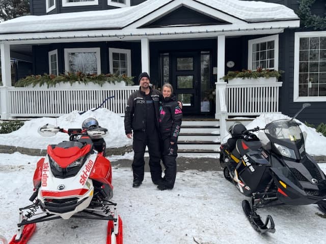 A man and a woman stand in front of a house, flanked by two snowmobiles on a snowy driveway. A man and a woman stand in front of a house, flanked by two snowmobiles on a snowy driveway.