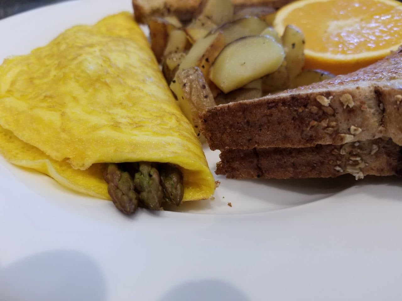 A plate featuring an asparagus omelette, toasted bread, sautéed potatoes, and a slice of orange.