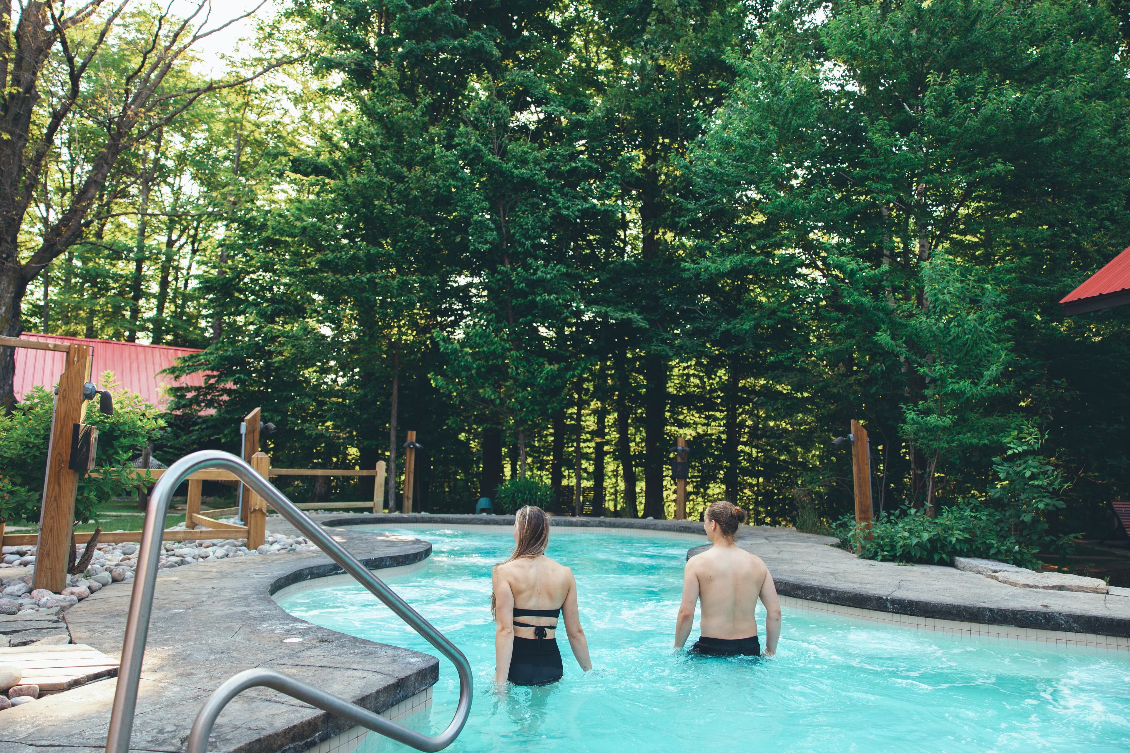 A couple walks through a serene outdoor pool surrounded by lush green trees.