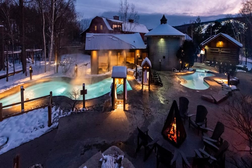 A serene winter resort featuring illuminated hot tubs and a fire pit surrounded by snow-covered landscapes.