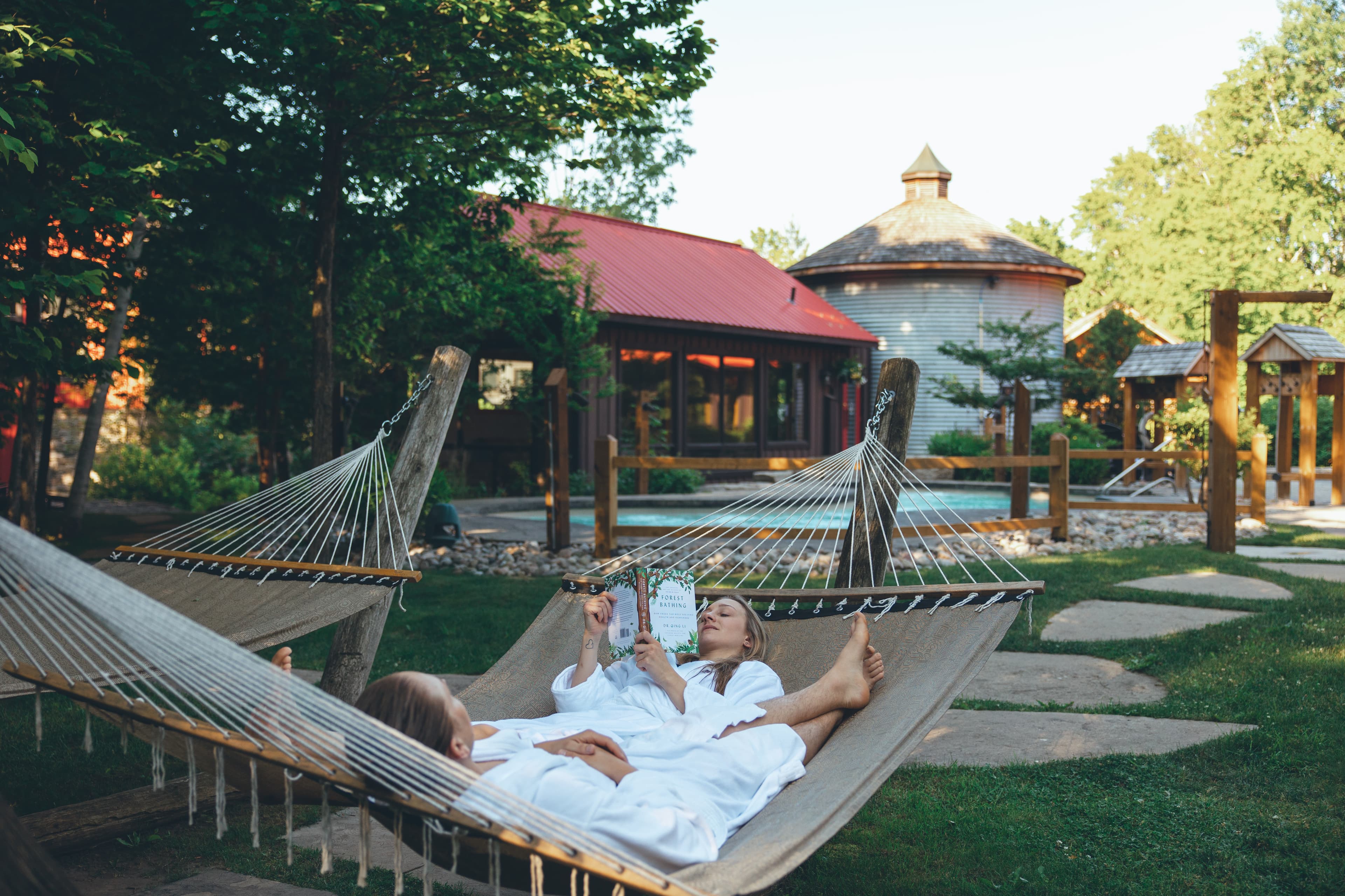 Two people in white robes relax in hammocks, surrounded by greenery and a rustic building.