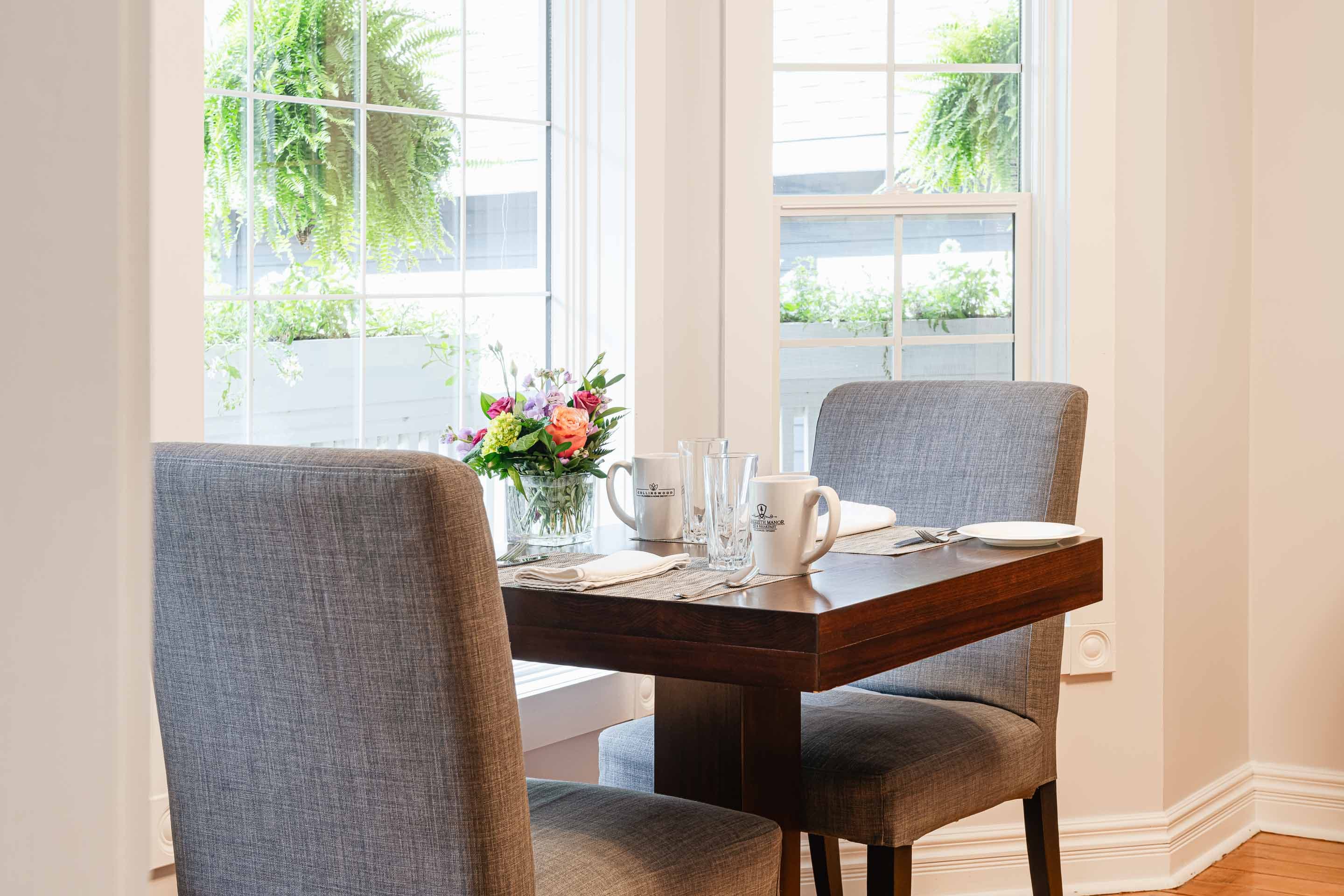 A cozy dining table set for two, adorned with a floral centerpiece and tableware, beside bright windows.