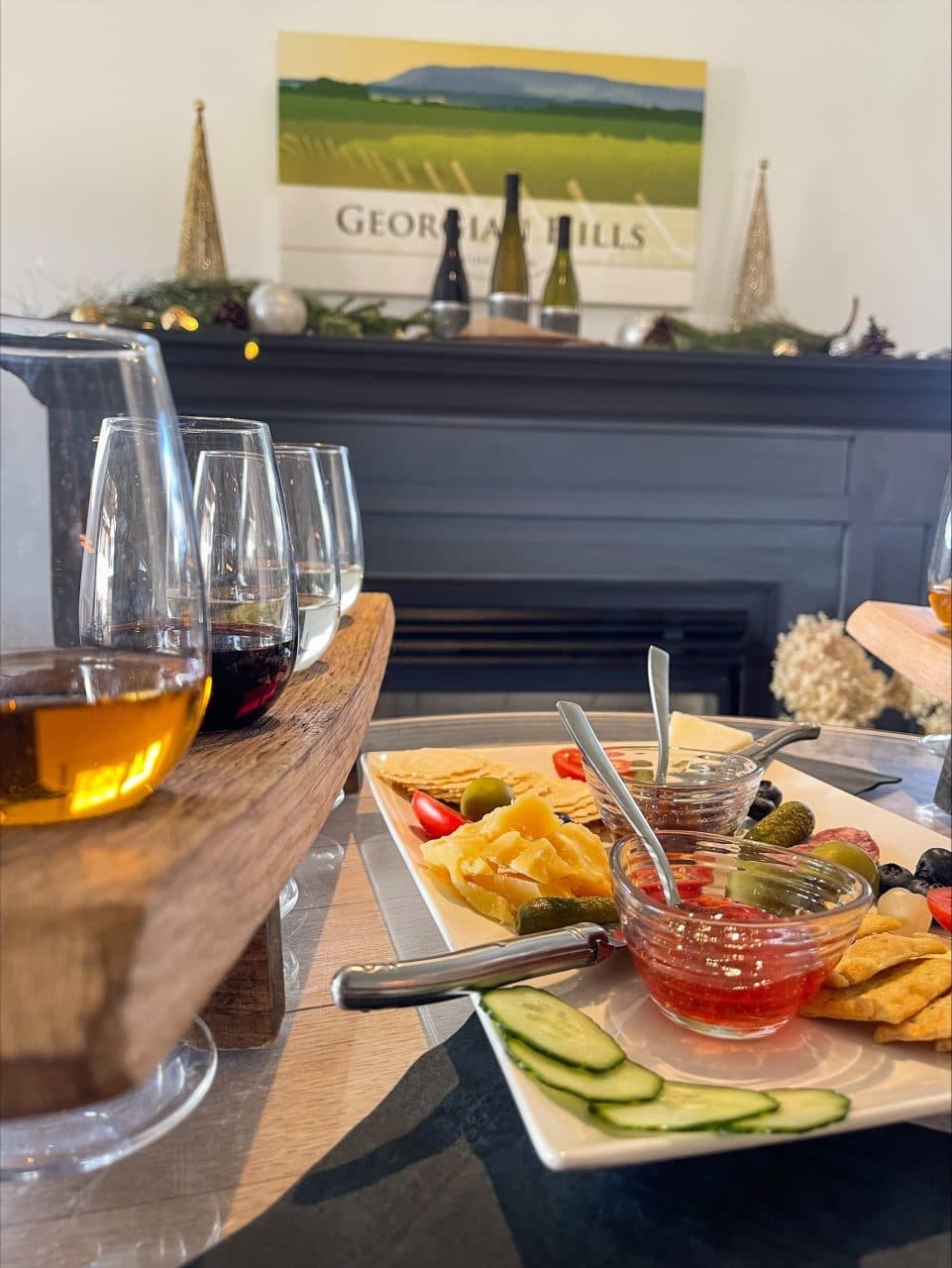 A wooden serving board with wine glasses and a platter of assorted cheeses, crackers, olives, and dips in a cozy setting.