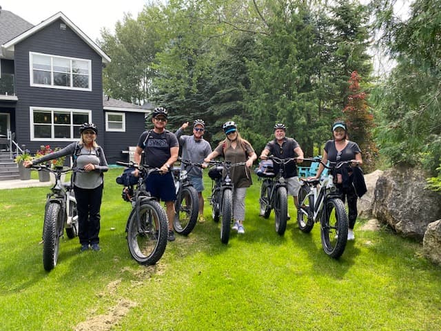 A group of six individuals poses with fat-tire bicycles in a grassy area outside a house.