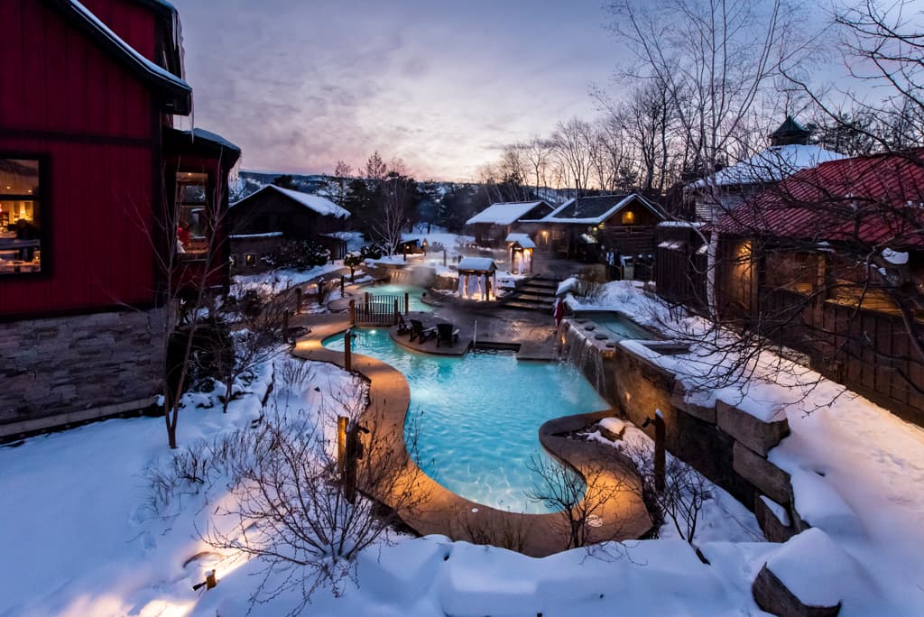 A snowy landscape features a lit outdoor pool and cozy cabins at dusk.