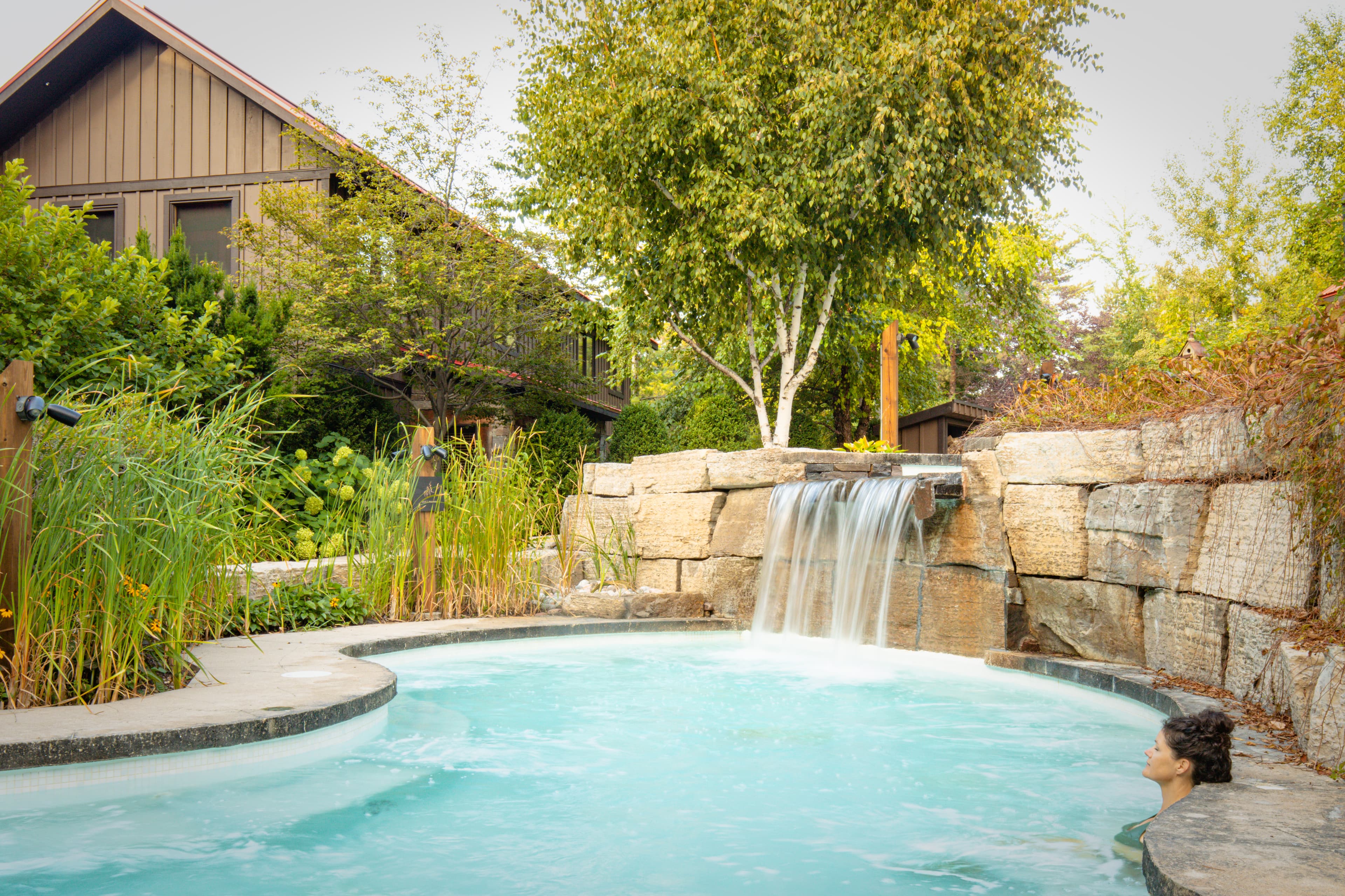 A serene hot tub with a waterfall surrounded by lush greenery and a rustic building in the background.