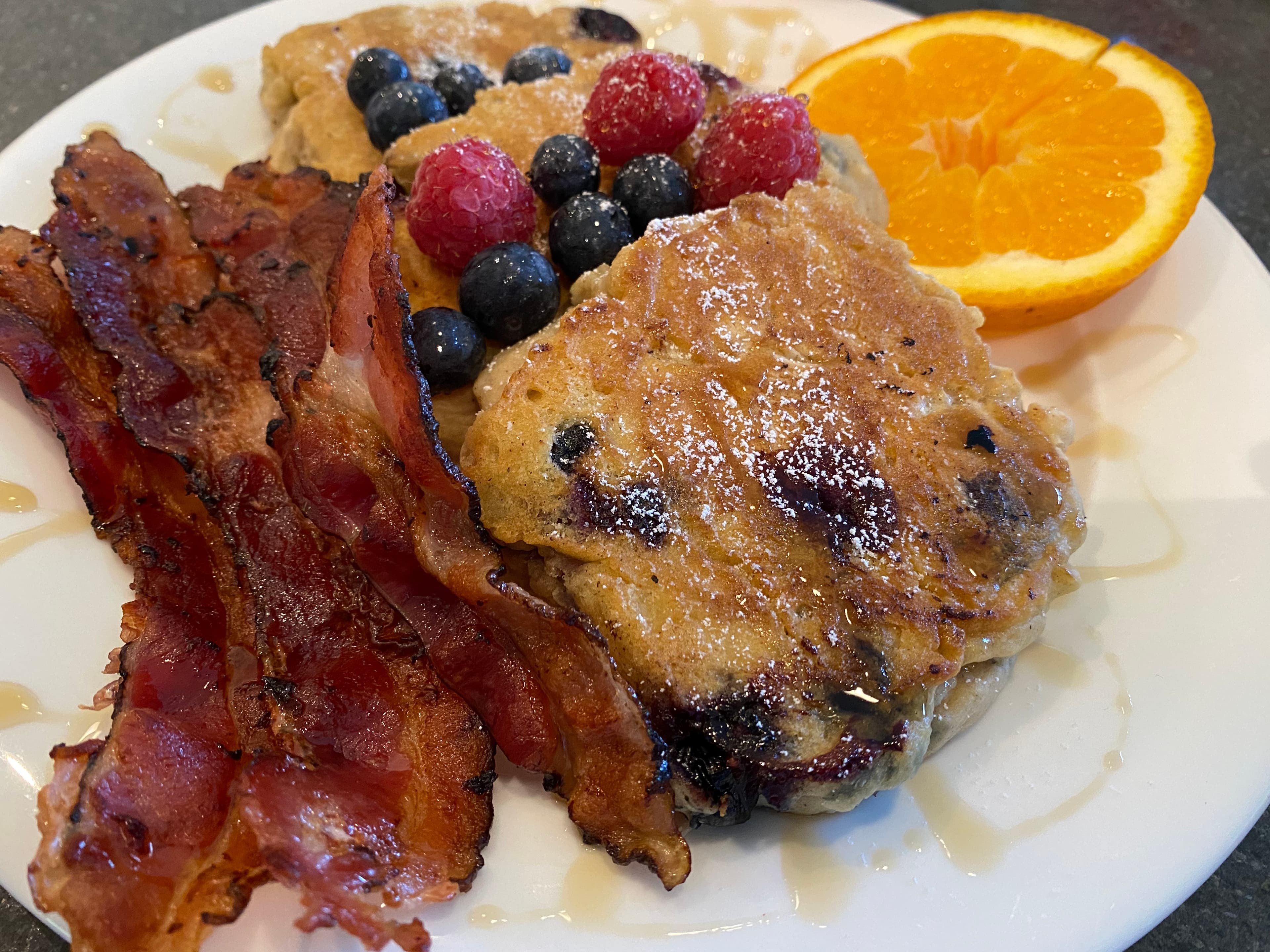 A plate of pancakes topped with berries, accompanied by crispy bacon and a slice of orange.