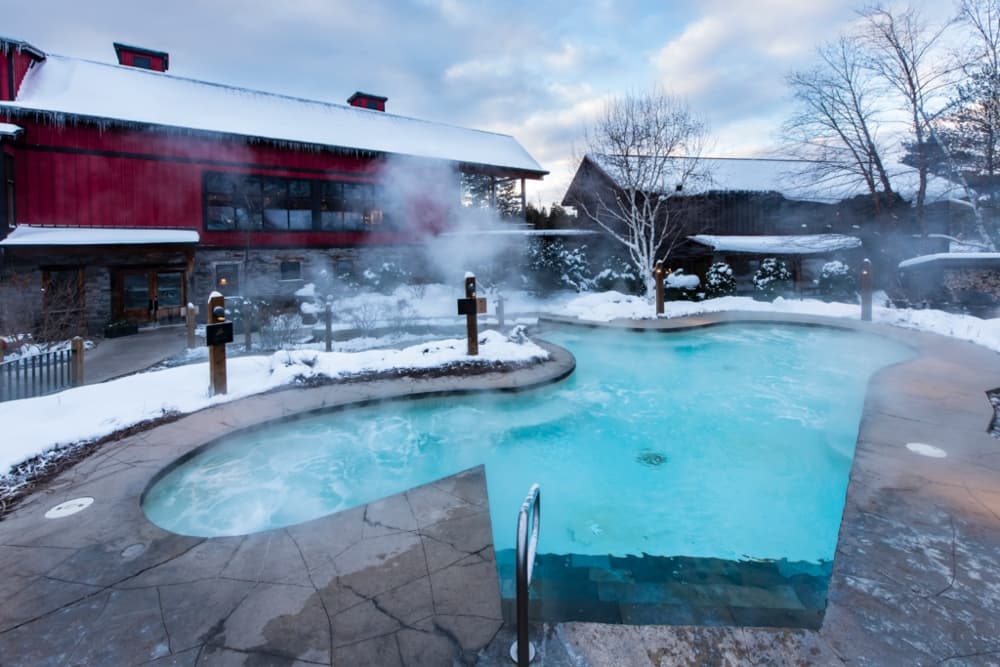 A steaming hot tub surrounded by snow and a cozy red building.